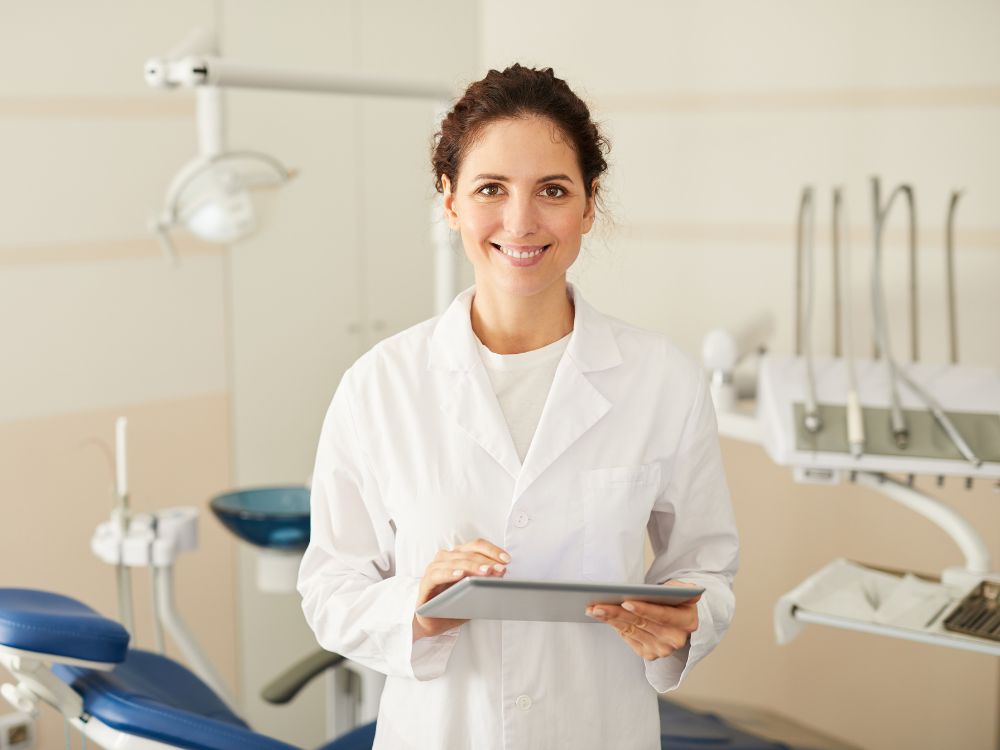 Dentista sorrindo em consultório odontológico segurando tablet, com cadeira e equipamentos ao fundo, ambiente limpo e profissional.