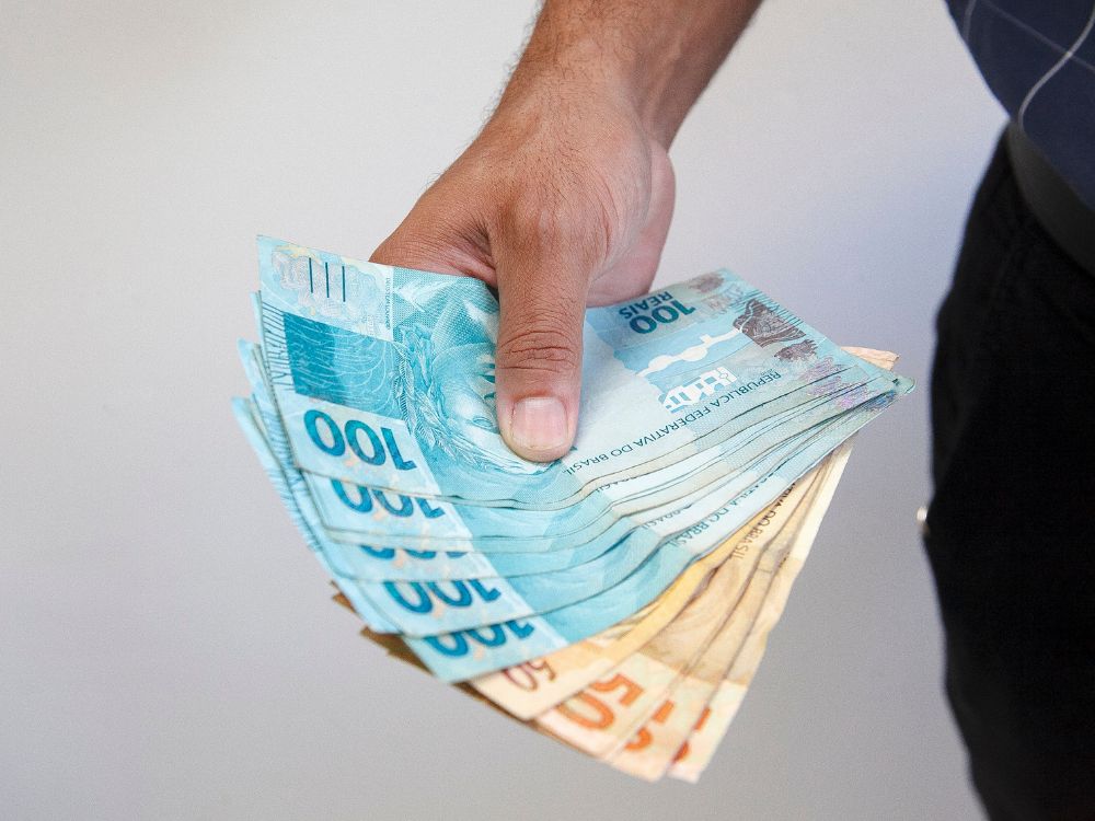 Mão segurando várias notas de real, incluindo cédulas de 100 e 50, representando dinheiro em espécie.