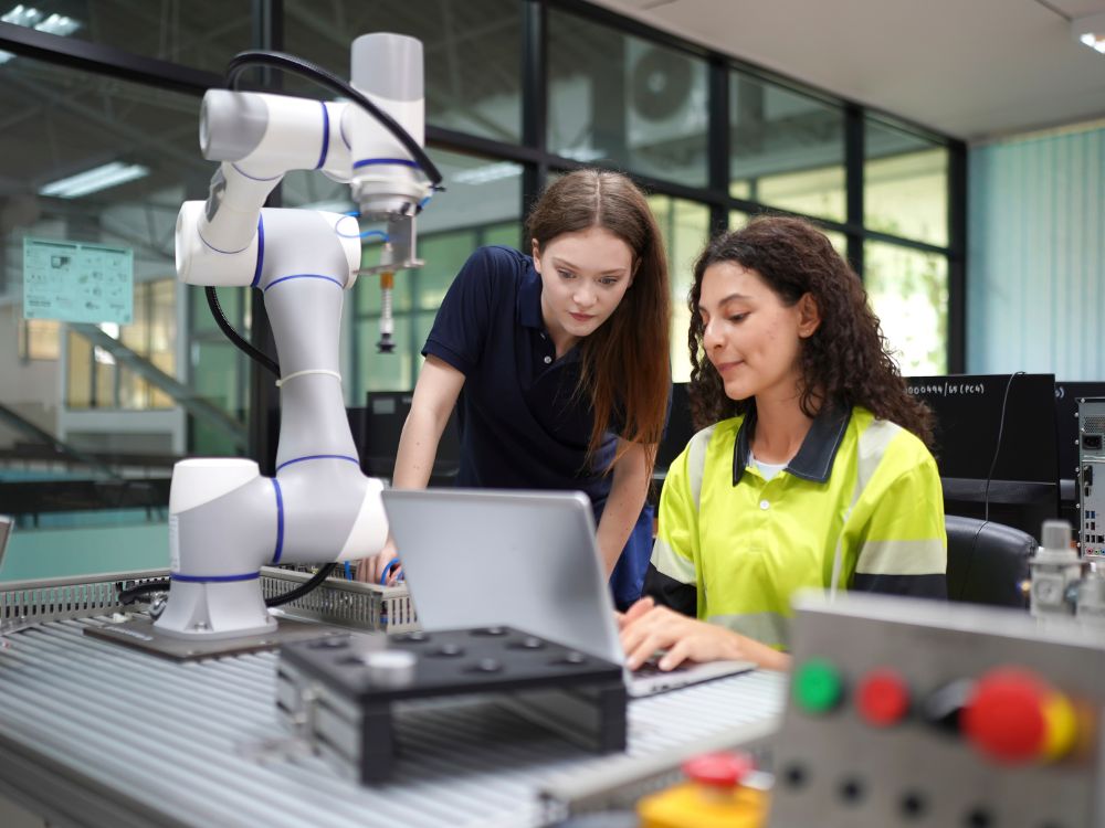 Texto alt: Estudantes trabalhando com robótica e tecnologia em laboratório industrial, utilizando notebook e braço robótico para atividades técnicas e automação.