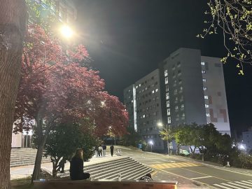 Área próxima à faculdade durante a noite, com prédios residenciais, iluminação urbana e estudantes circulando, destacando a rotina no entorno universitário.