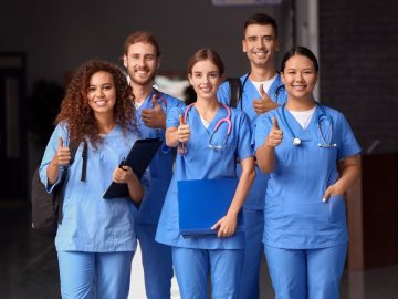 Equipe de profissionais de enfermagem com uniformes azuis e estetoscópios, sorrindo e fazendo sinal de positivo, representando união e dedicação na área da saúde.